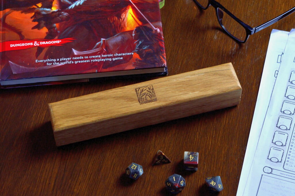 An Oak Artisan Dice Vault with dice, rule book and character sheets. There are also some glasses on the tabletop.